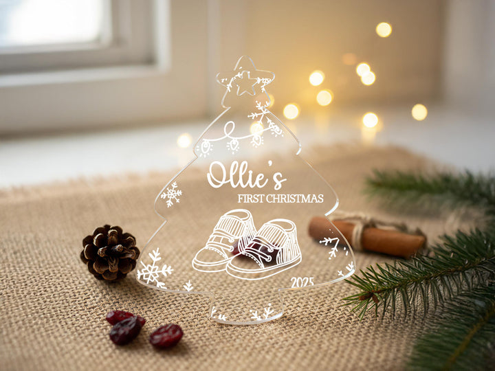 A clear glass ornament shaped like a Christmas tree with a snowflake design, featuring the text &quot;Ollie&#39;s FIRST CHRISTMAS&quot; and a pair of baby shoes. The ornament is placed on a burlap surface, surrounded by pine branches, cranberries, and a pine cone.