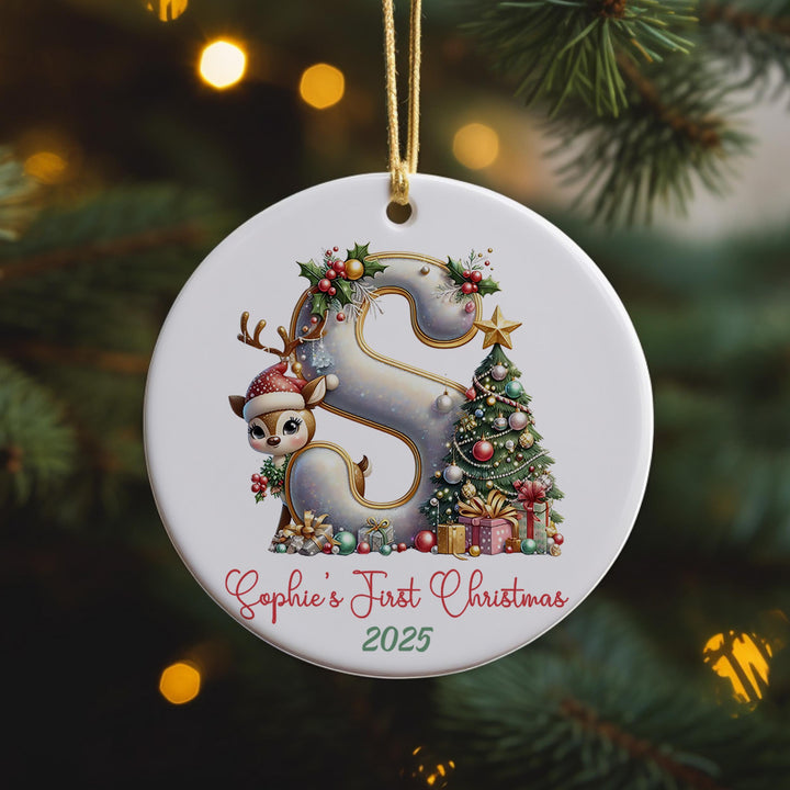 A white ornament with a decorative letter &#39;S&#39; and a small reindeer, surrounded by Christmas decorations, hangs on a tree.