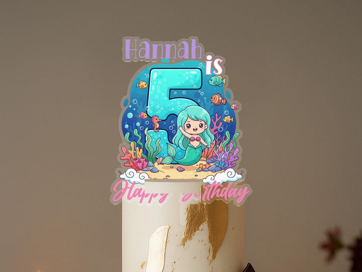 A birthday cake with a mermaid-themed topper, featuring the name &quot;Hannah&quot; and the number 5.