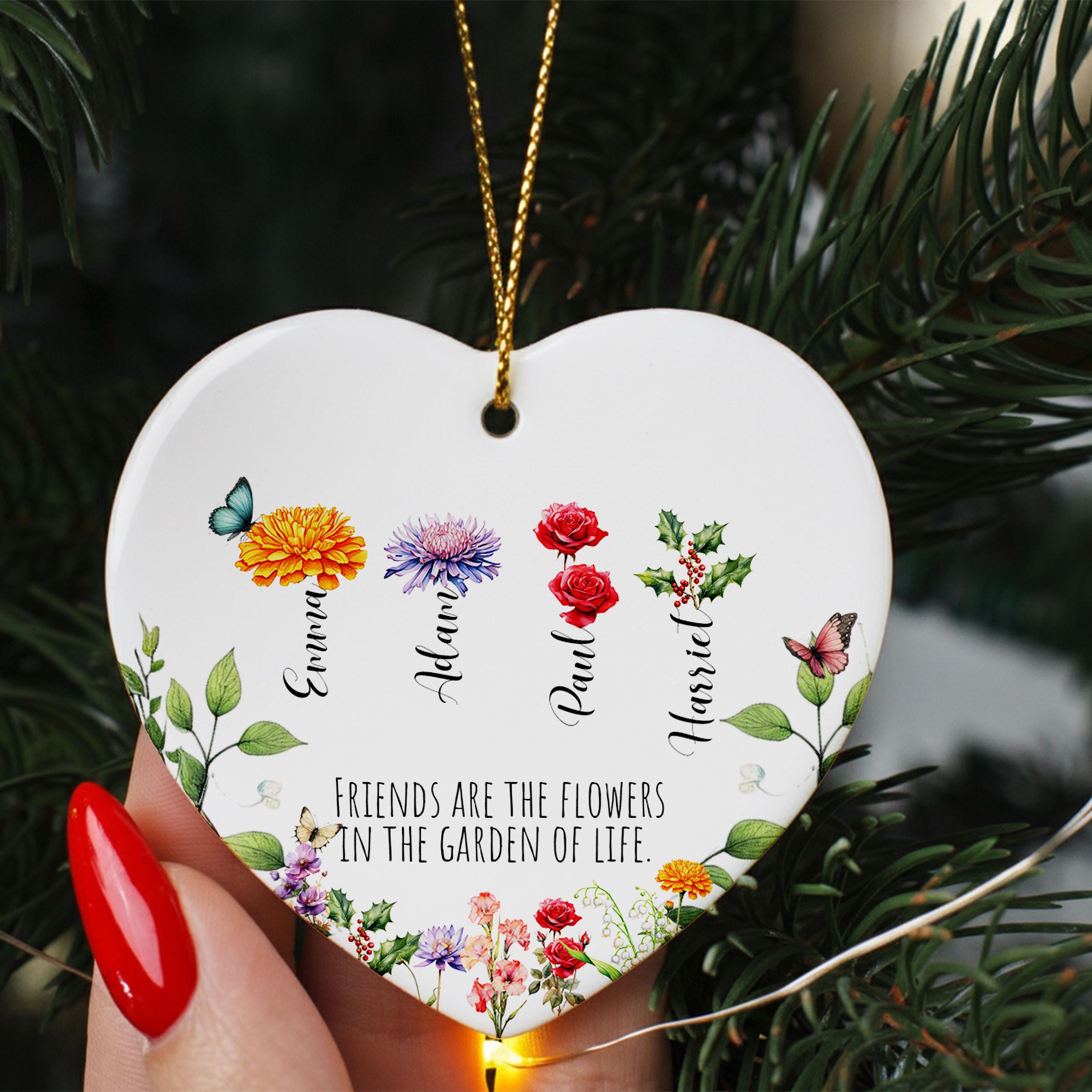 A heart-shaped ornament with a floral design and the text "Friends are the flowers in the garden of life" is being held in front of a pine tree.