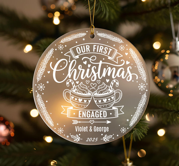 A Christmas ornament with the text &quot;Our first Christmas&quot; and &quot;Engaged&quot; on it, along with illustrations of cups and snowflakes.