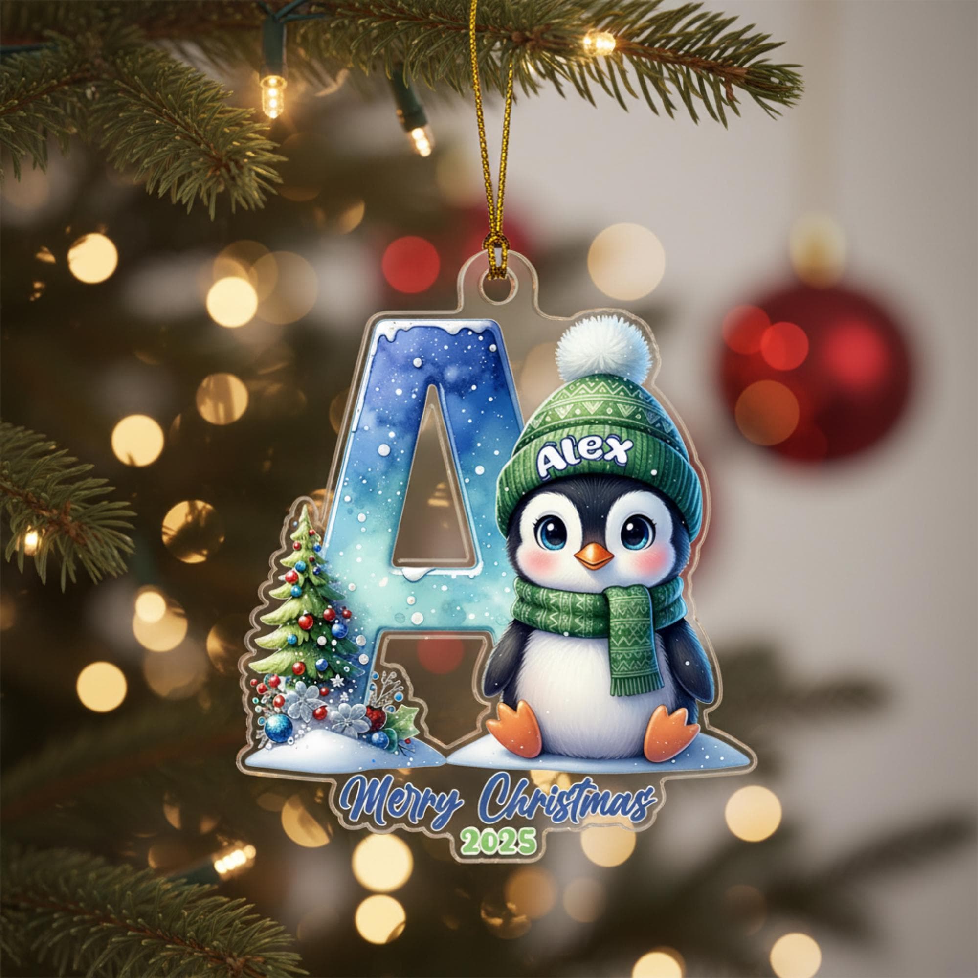 A Christmas ornament featuring a penguin wearing a green hat and scarf, with the letter "A" and a snowflake design, hanging on a tree.