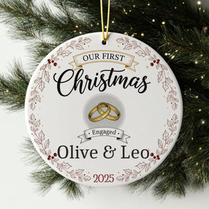 A white Christmas ornament with the text "Our first Christmas" and "Engaged Olive & Leo" printed on it, along with a graphic of a ring and holly leaves.