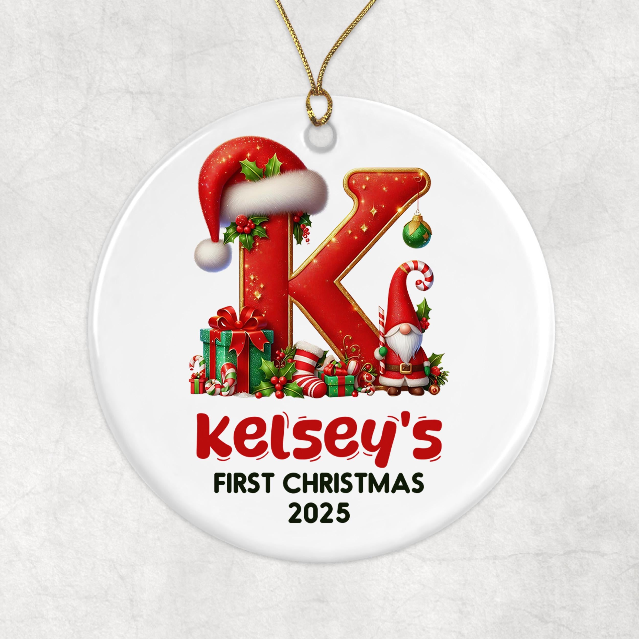 A white oval ornament with a red letter "K" and various Christmas-themed elements, including a Santa hat, presents, and a gnome. The ornament is labeled "Kelsey's First Christmas 2020".