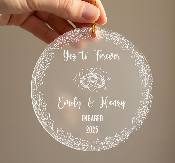Personalised engagement Christmas ornament held in hand showing frosted acrylic design with Yes to Forever text, wedding rings with hearts and sparkles, couple names Emily and Henry, Engaged 2025, surrounded by holly leaf and berry border with gold ribbonRetry