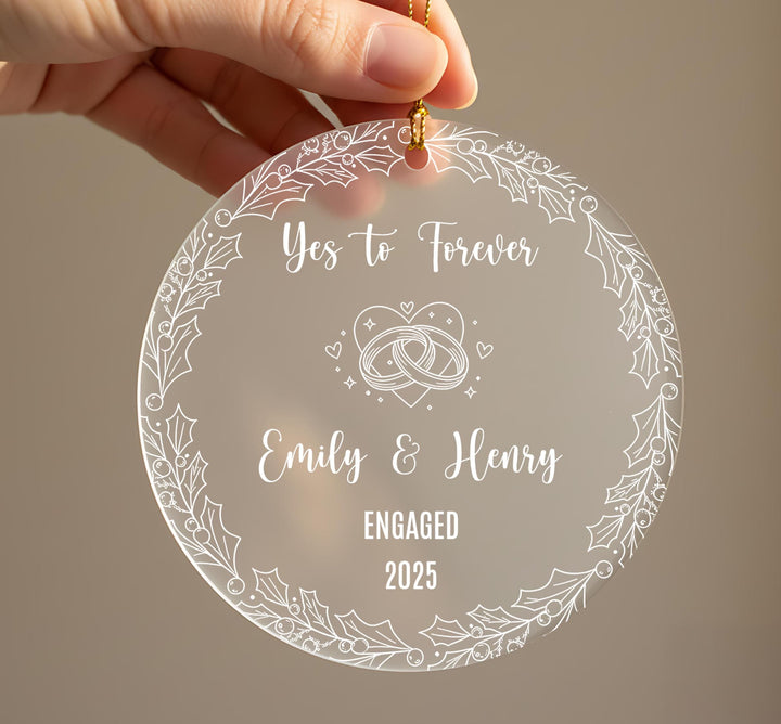 Personalised engagement Christmas ornament held in hand showing frosted acrylic design with Yes to Forever text, wedding rings with hearts and sparkles, couple names Emily and Henry, Engaged 2025, surrounded by holly leaf and berry border with gold ribbonRetry