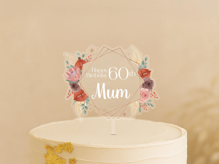 A white cake with a clear floral topper that says &quot;Happy Birthday 60th Mom&quot; in a decorative font.