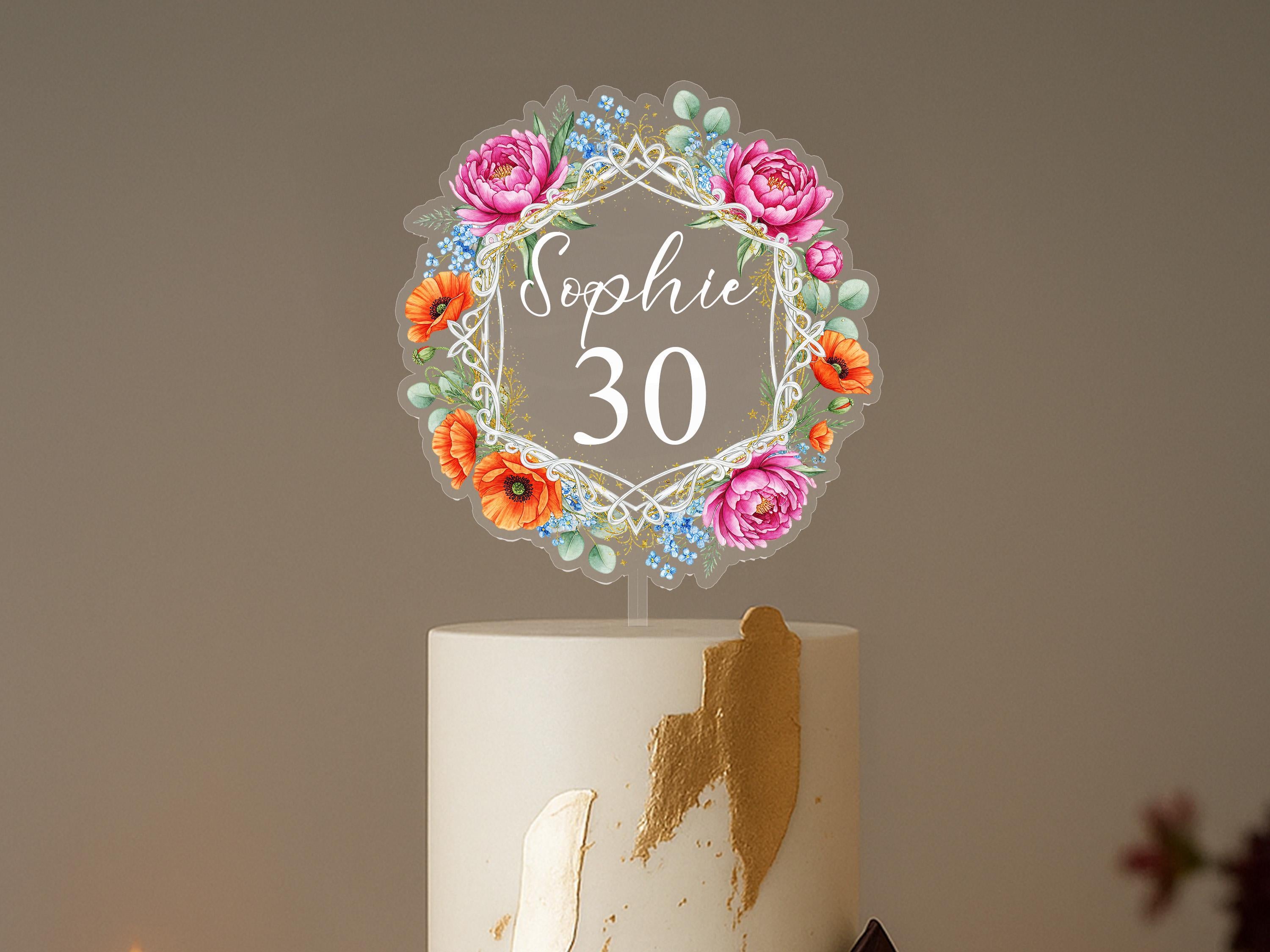 A cake topper with a floral design and the name "Sophie" written on it, placed on top of a white cake.