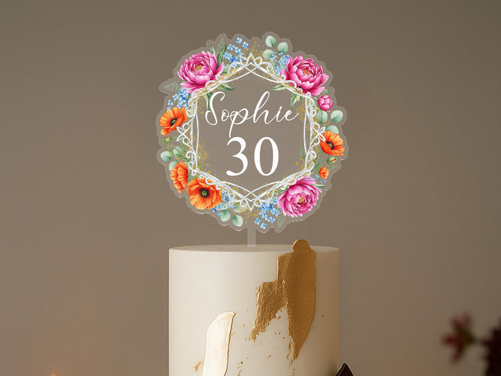 A cake topper with a floral design and the name &quot;Sophie&quot; written on it, placed on top of a white cake.