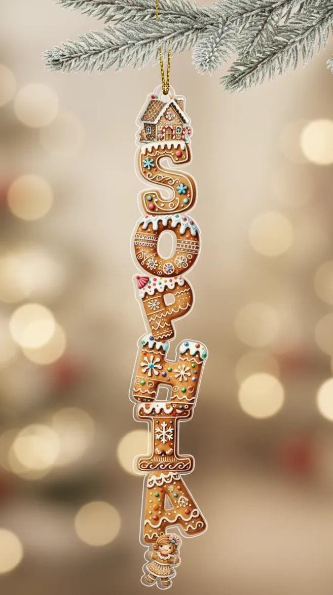A gingerbread house ornament with the name "Sophia" made up of gingerbread letters hanging from a branch of a pine tree.