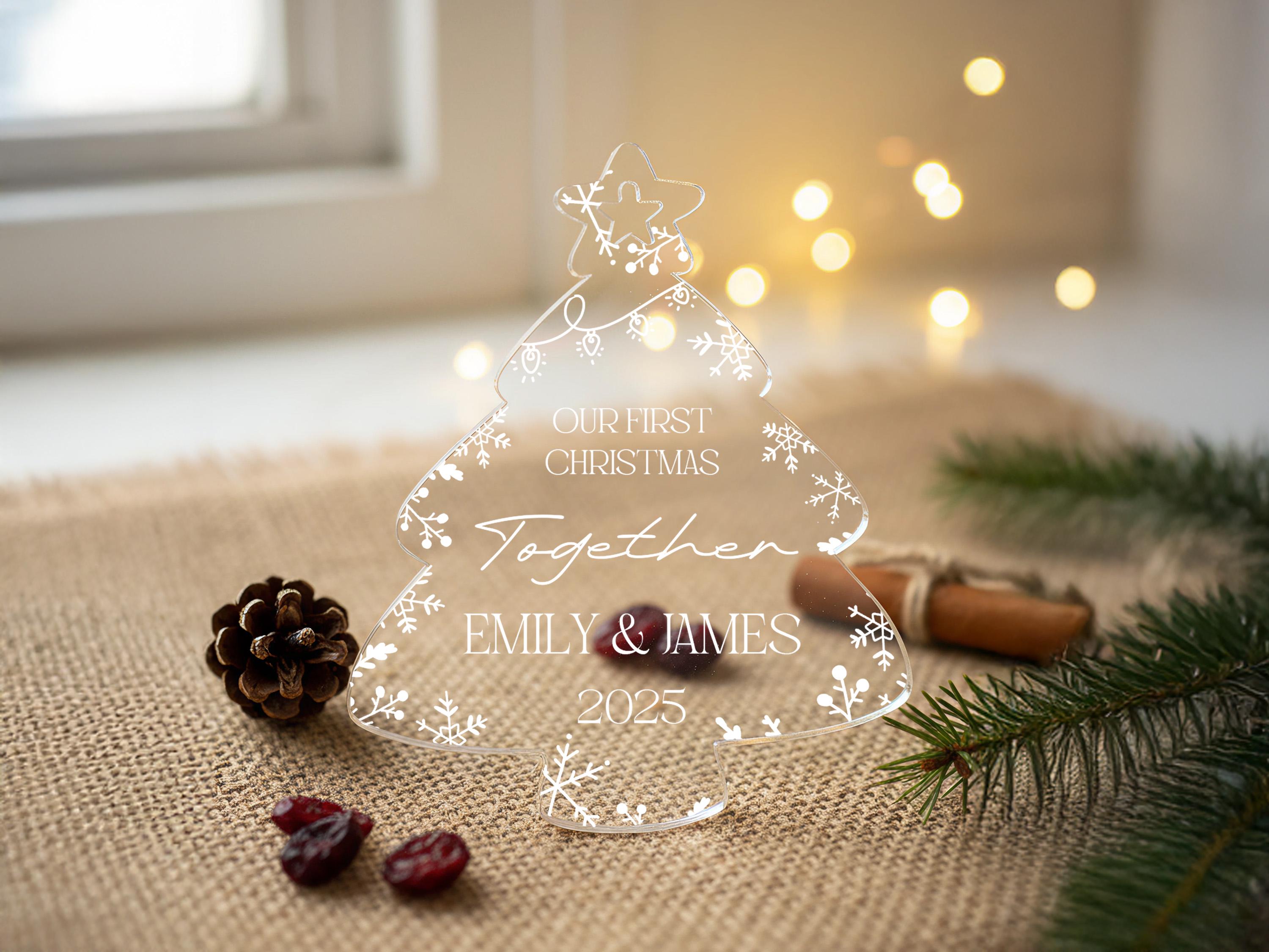 A transparent Christmas tree-shaped object with the text "Our first Christmas together" and "EMILY & JAMES" written on it, surrounded by pine branches, cranberries, and a star ornament.