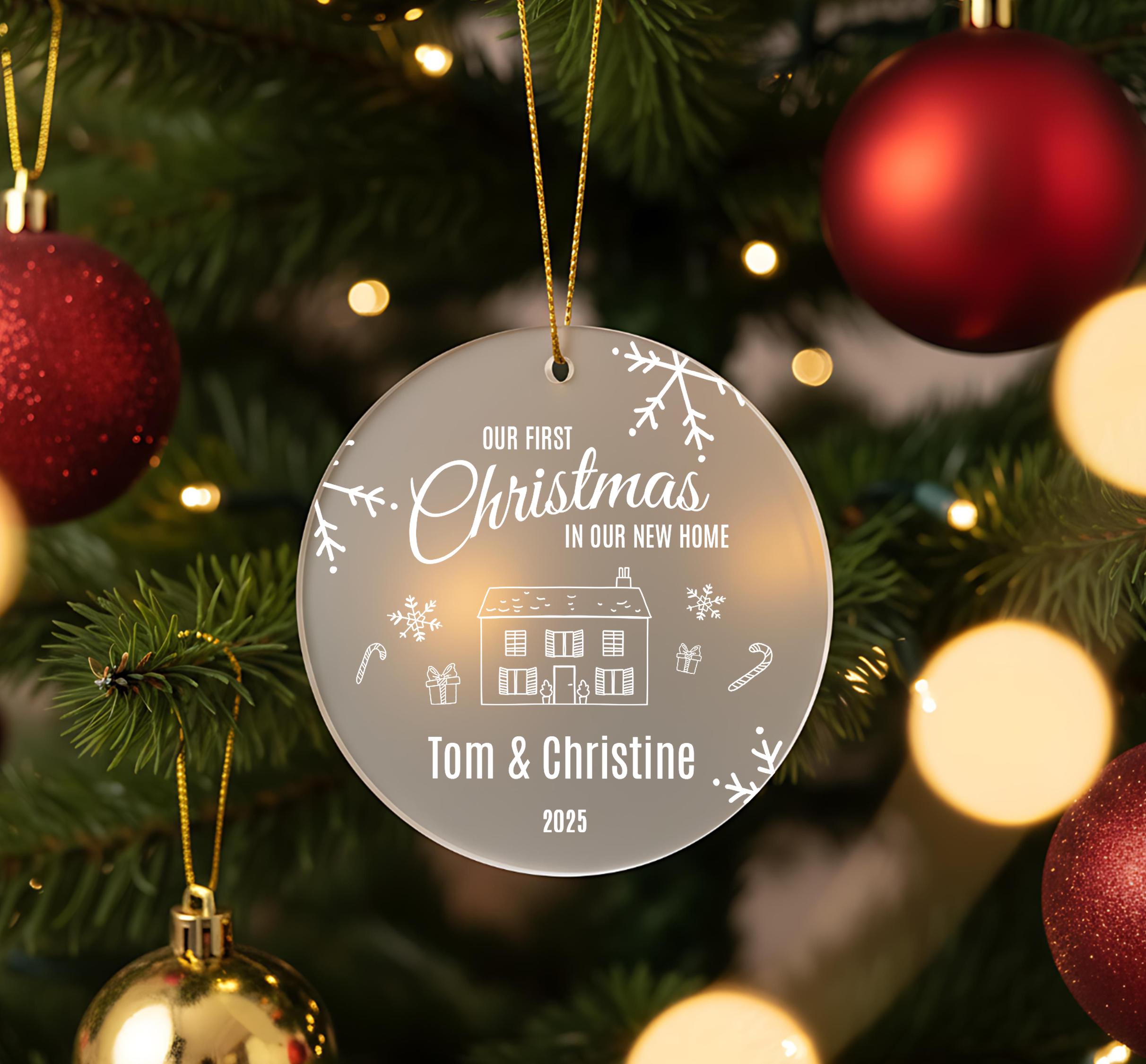 A clear ornament hanging on a Christmas tree, featuring a house and snowflakes, with the text "Our first Christmas in our new home" and the names "Tom & Christine" and "2023" below it.