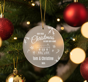 A clear ornament hanging on a Christmas tree, featuring a house and snowflakes, with the text "Our first Christmas in our new home" and the names "Tom & Christine" and "2023" below it.