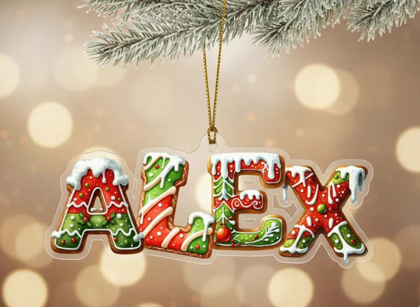 A clear ornament shaped like the name &quot;ALEX&quot; made of colorful gingerbread cookies, hanging from a branch with a blurred background of lights.