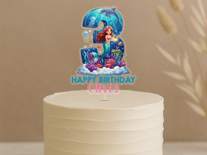 Personalised mermaid birthday cake topper featuring number 3 with red-haired mermaid, dolphin, jellyfish, sea turtle, coral reef and custom name Olivia on white celebration cake