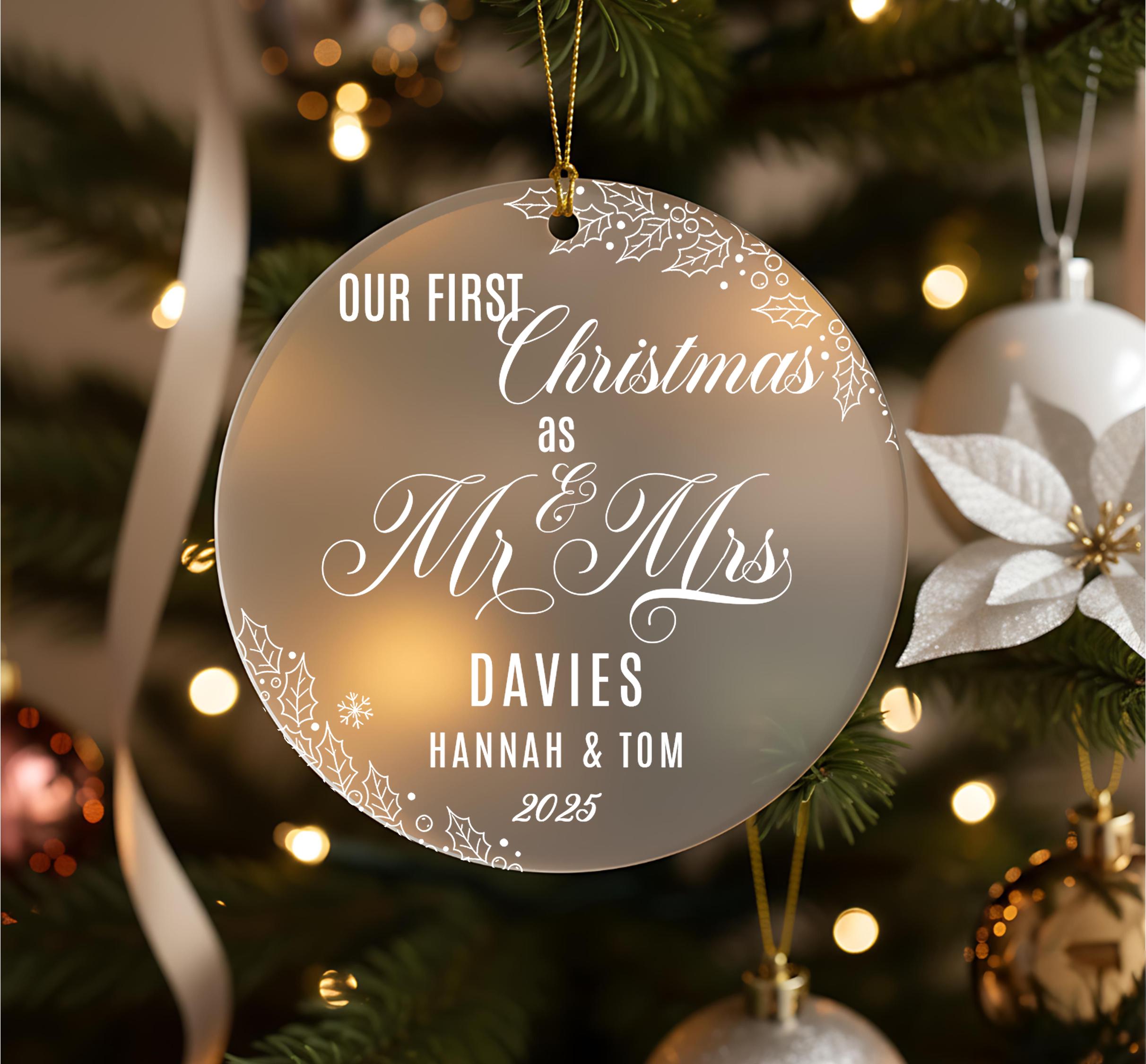 A Christmas ornament with the text "OUR FIRST Christmas as Mr & Mrs DAVIES Hannah & Tom 2035" on it, hanging on a tree with other ornaments and lights visible in the background.