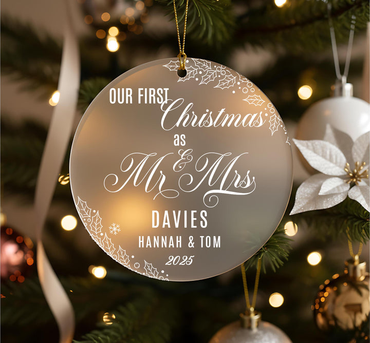A Christmas ornament with the text &quot;OUR FIRST Christmas as Mr & Mrs DAVIES Hannah & Tom 2035&quot; on it, hanging on a tree with other ornaments and lights visible in the background.