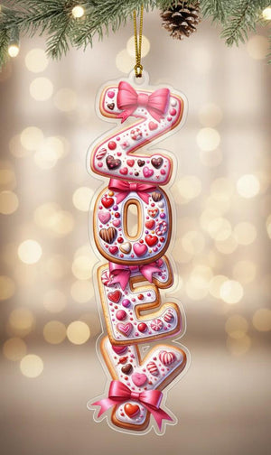 A clear ornament shaped like the name "Zoey" made of colorful candy pieces and bows, hanging on a branch with Christmas lights in the background.