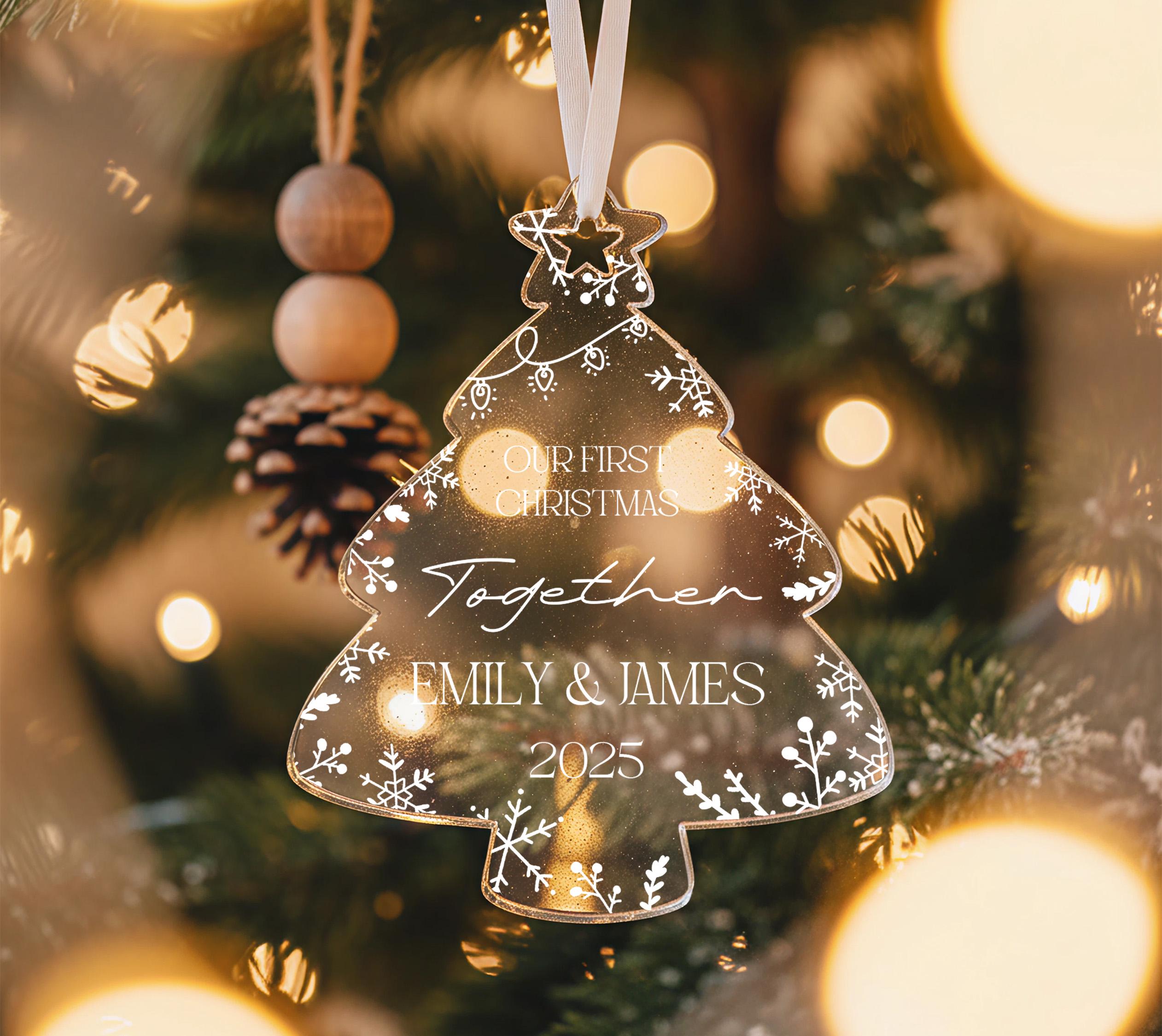 A clear glass ornament shaped like a Christmas tree hangs on a tree, with a pine cone ornament hanging from it. The ornament has a message that reads "Our first Christmas together Emily & James 2015".