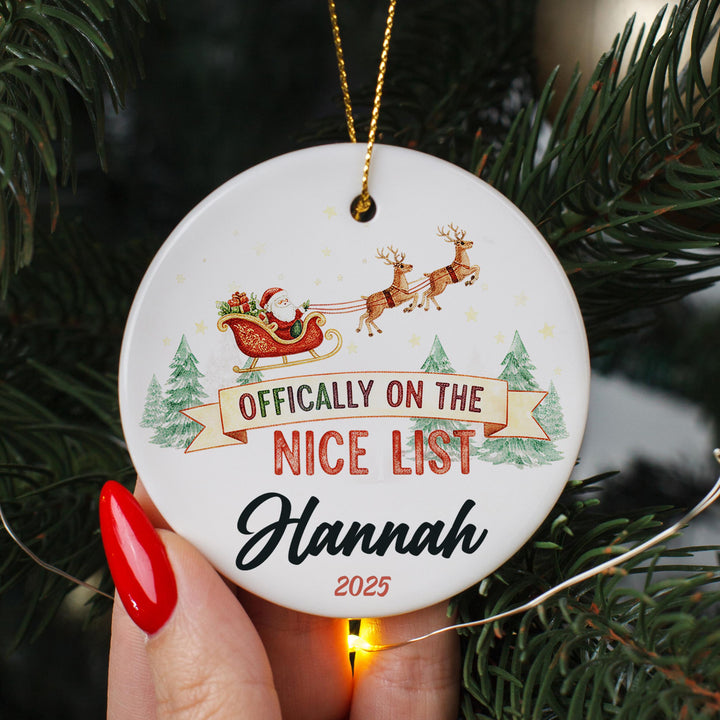 A hand holding a white ornament with a Santa Claus sleigh and reindeer, along with the text &quot; Officially on the NICE LIST Hannah 2020&quot;.