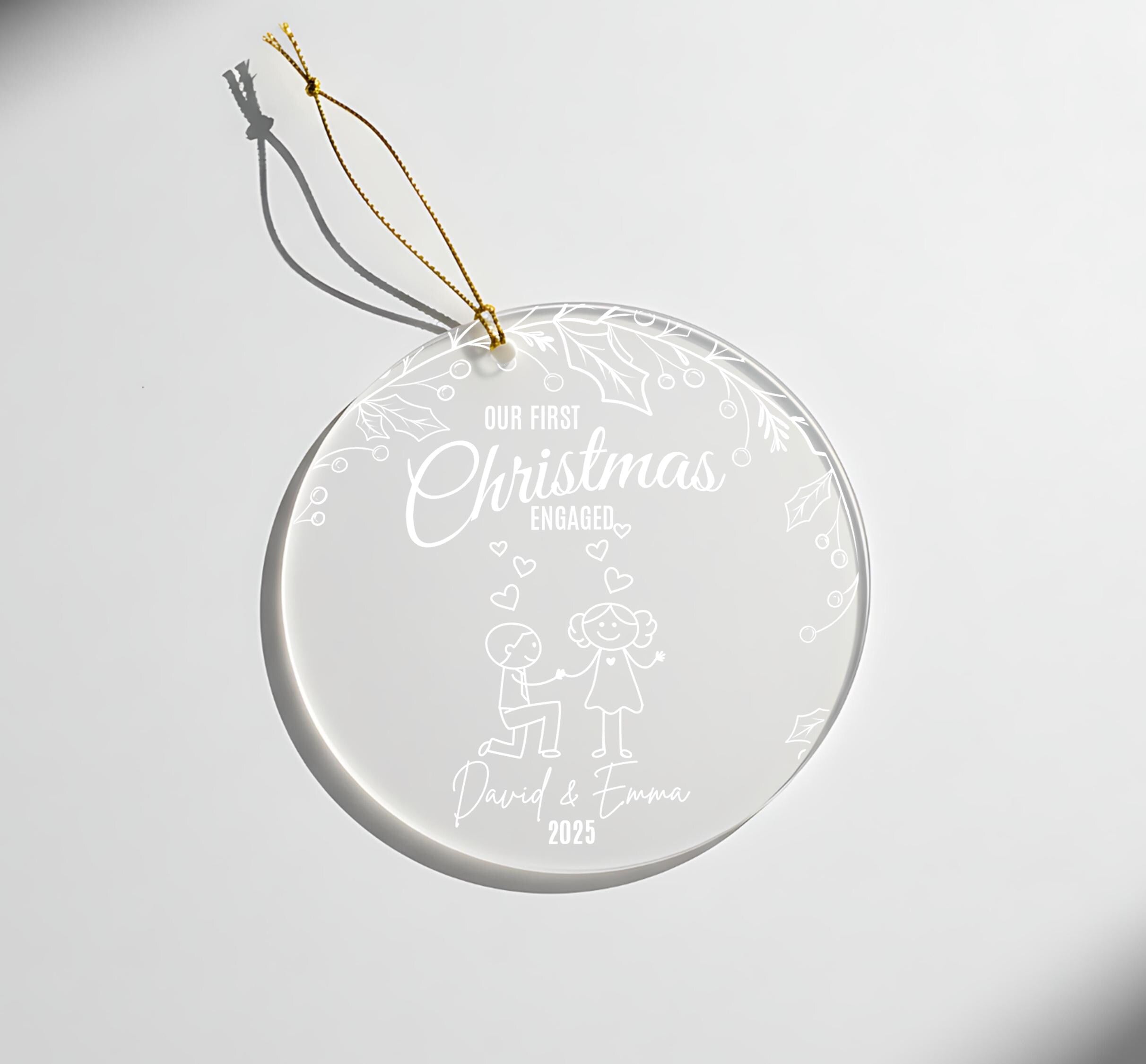 A clear glass ornament with a gold string attached, featuring a design of a couple and the text "Our first Christmas engaged David & Emma 2015".