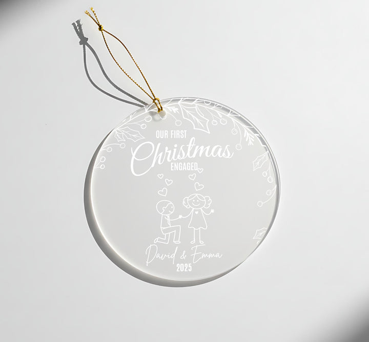 A clear glass ornament with a gold string attached, featuring a design of a couple and the text &quot;Our first Christmas engaged David & Emma 2015&quot;.