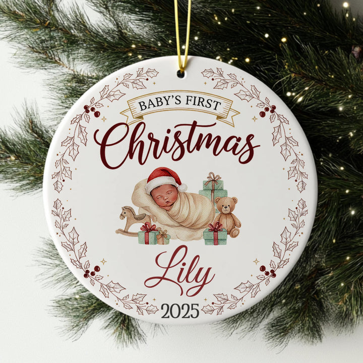 A white ornament with a baby and Christmas-themed decorations, hanging on a pine tree branch.
