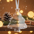 A clear ornament shaped like a Christmas tree with the text "Our first Christmas together" and "EMILY & JAMES" printed on it, hanging from a ribbon. The ornament is surrounded by pine cones, cinnamon sticks, and greenery, with a blurred background of lights and snow.