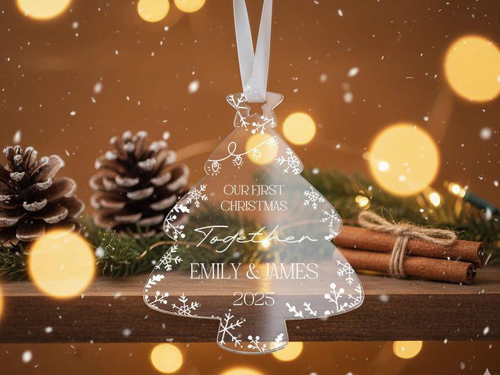 A clear ornament shaped like a Christmas tree with the text &quot;Our first Christmas together&quot; and &quot;EMILY & JAMES&quot; printed on it, hanging from a ribbon. The ornament is surrounded by pine cones, cinnamon sticks, and greenery, with a blurred background of lights and snow.