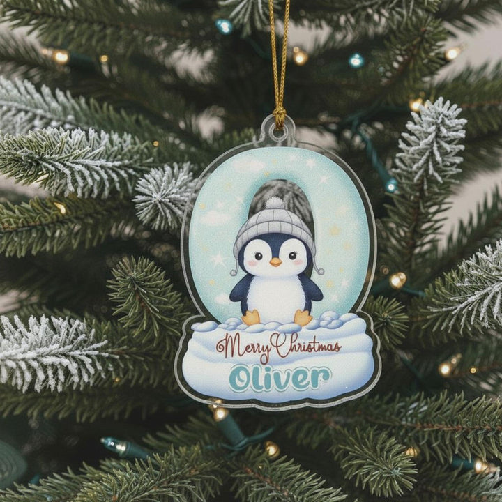 A clear ornament with a cartoon penguin wearing a hat and sitting on a snowy surface, surrounded by Christmas lights on a decorated tree.