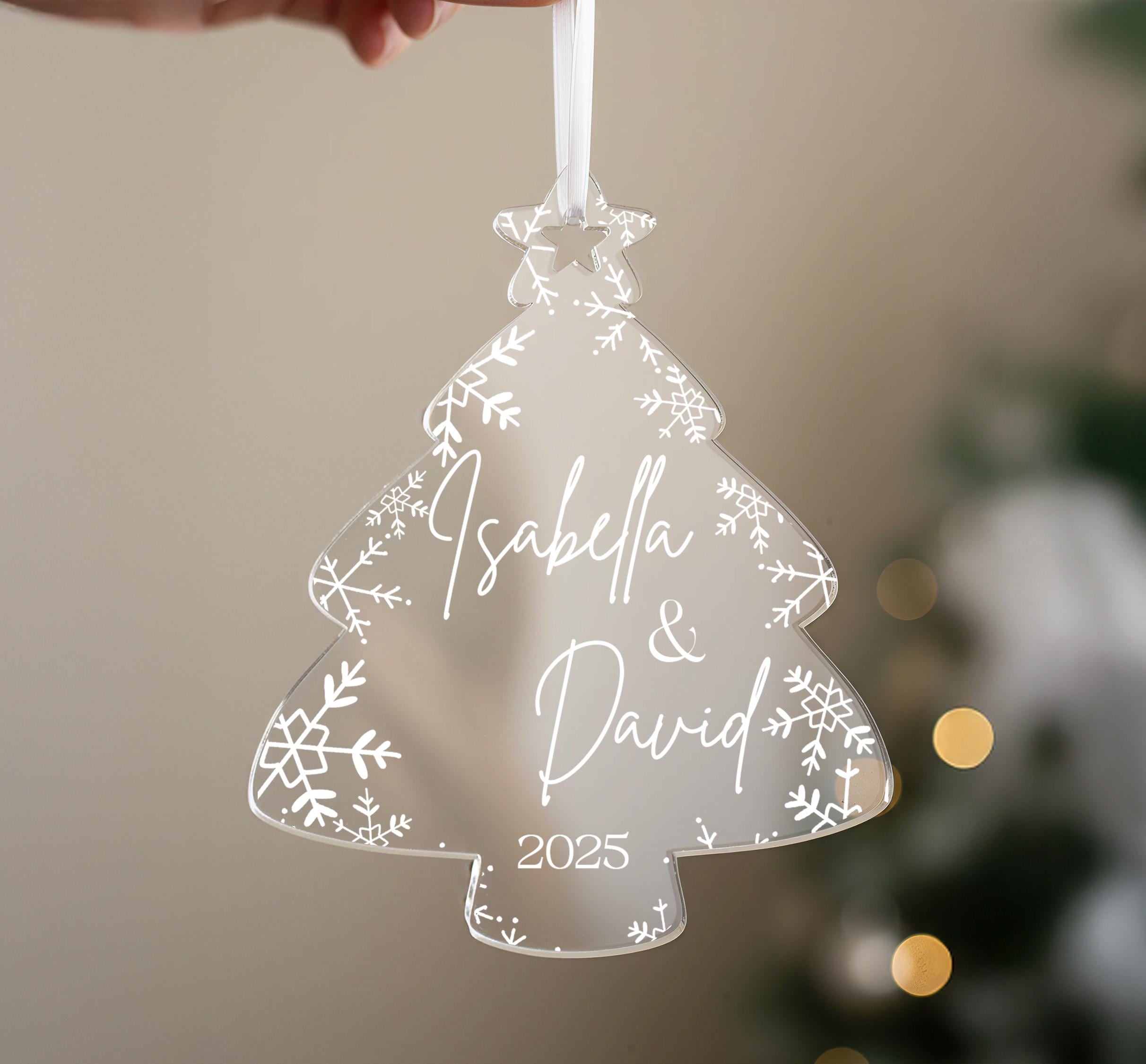 A hand holding a transparent ornament shaped like a Christmas tree with snowflakes and the names "Isabella & David" written on it.
