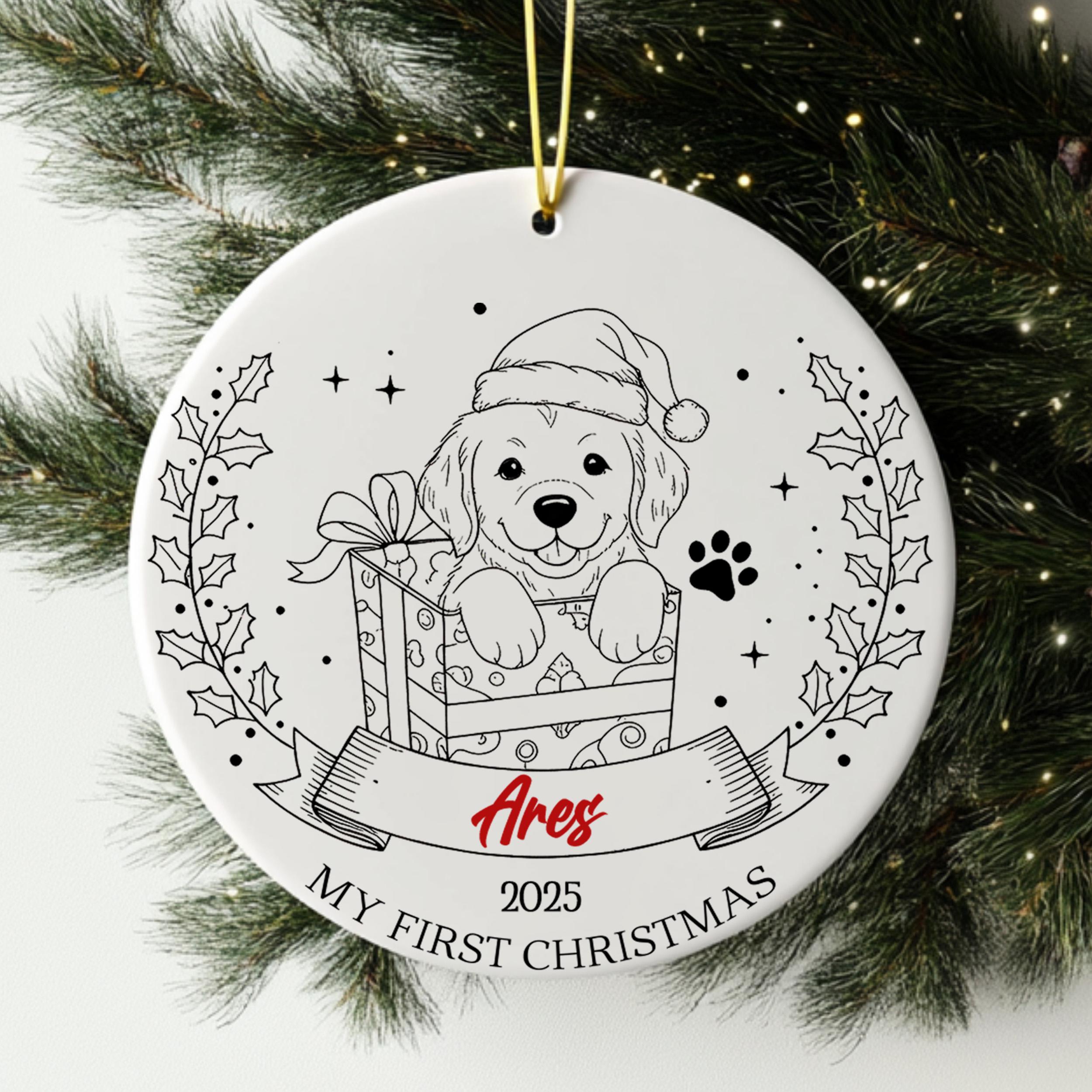 A white ceramic ornament with a black and white illustration of a dog wearing a Santa hat, holding a gift box, and surrounded by holly leaves and a banner with the text "Ares" and "My first Christmas".