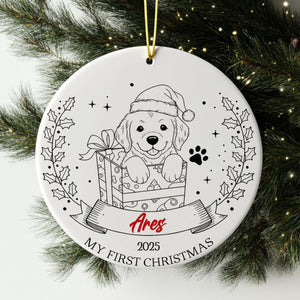 A white ceramic ornament with a black and white illustration of a dog wearing a Santa hat, holding a gift box, and surrounded by holly leaves and a banner with the text "Ares" and "My first Christmas".