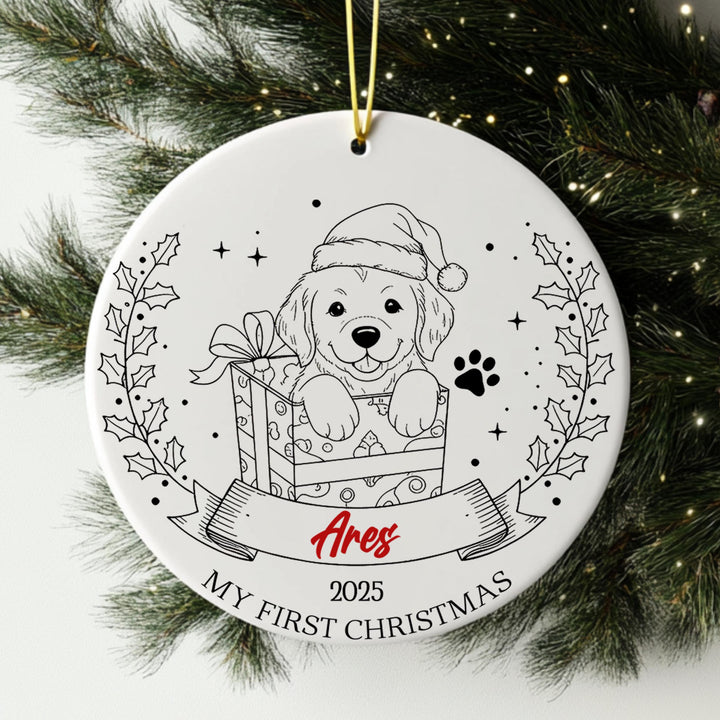 A white ceramic ornament with a black and white illustration of a dog wearing a Santa hat, holding a gift box, and surrounded by holly leaves and a banner with the text &quot;Ares&quot; and &quot;My first Christmas&quot;.