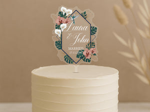A white wedding cake with a decorative topper featuring the names "Laura & John" and the date "17.11.26" surrounded by floral elements.