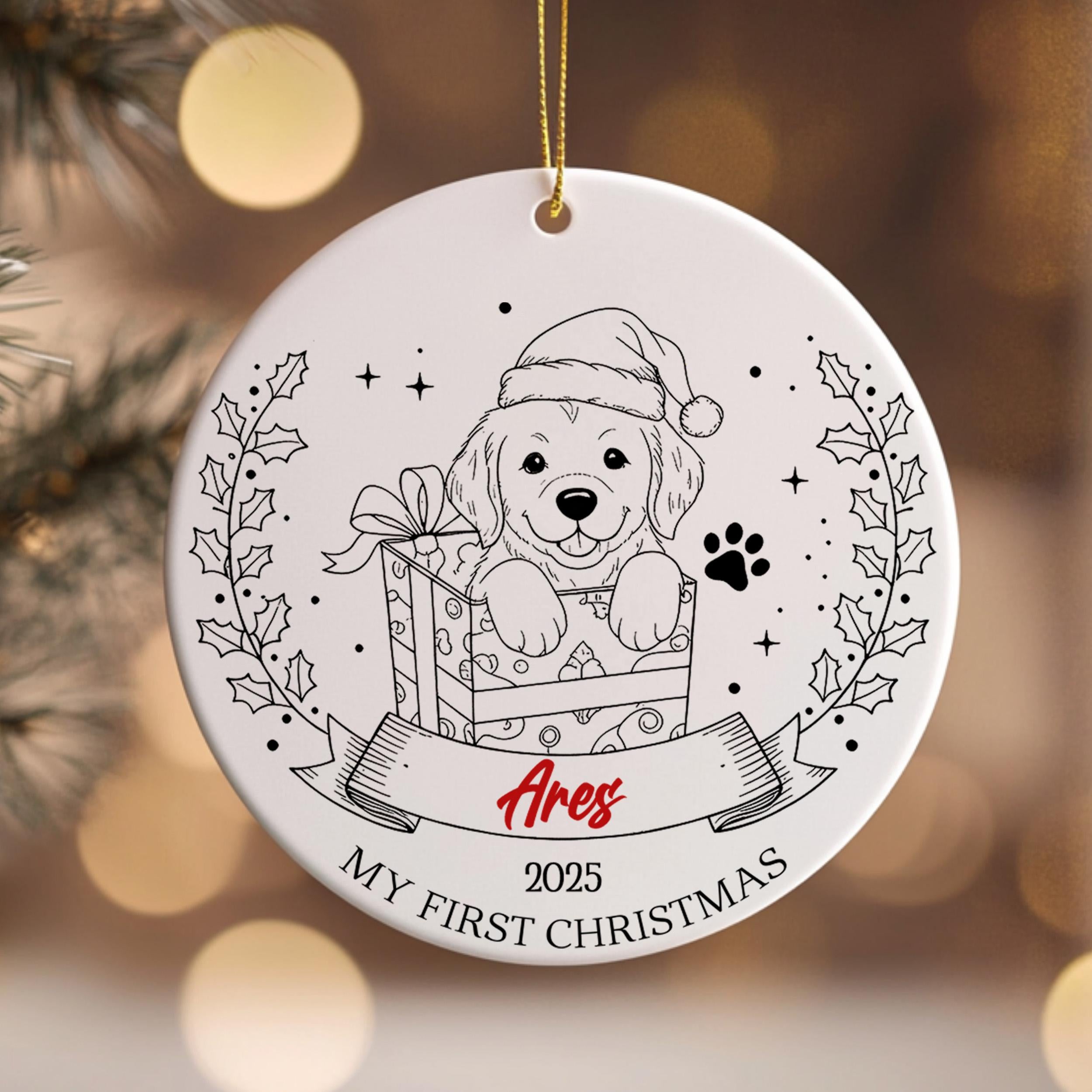 A white ceramic ornament with a black and white illustration of a dog wearing a Santa hat, holding a gift box, and surrounded by holly leaves and a banner with the text "Ares" and "My first Christmas".