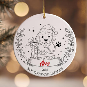 A white ceramic ornament with a black and white illustration of a dog wearing a Santa hat, holding a gift box, and surrounded by holly leaves and a banner with the text "Ares" and "My first Christmas".