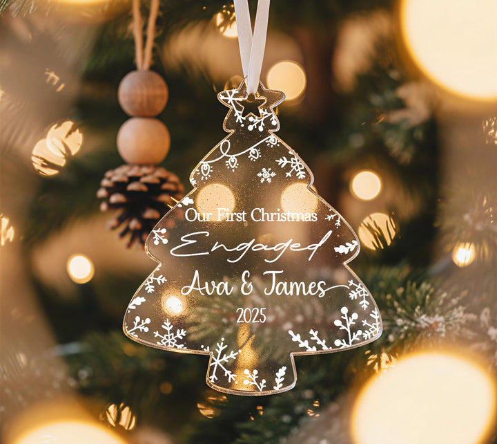 A clear Christmas ornament shaped like a tree hangs on a tree, with a personalized message engraved on it.
