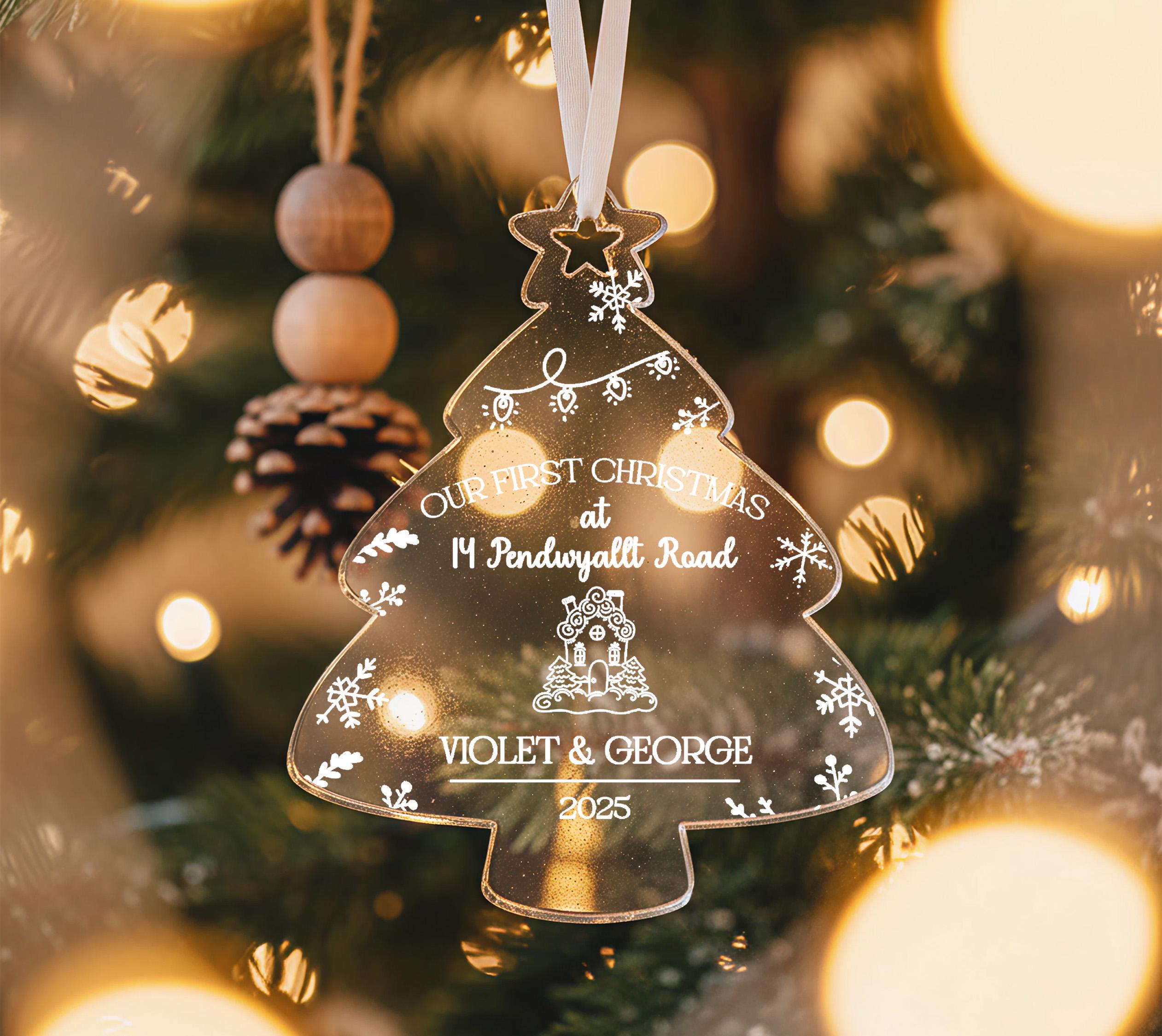 A clear glass ornament shaped like a Christmas tree hangs on a tree, with a personalized message about the couple's first Christmas at 14 Pendyvalt Road.