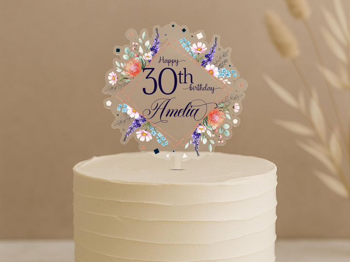 A white cake with a decorative floral topper that says "Happy 30th birthday Amelia".