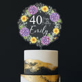 A birthday cake with a wreath-shaped topper that says "Happy Birthday Emily" and features purple and yellow flowers.