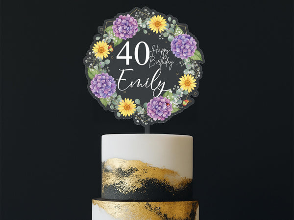 A birthday cake with a wreath-shaped topper that says &quot;Happy Birthday Emily&quot; and features purple and yellow flowers.