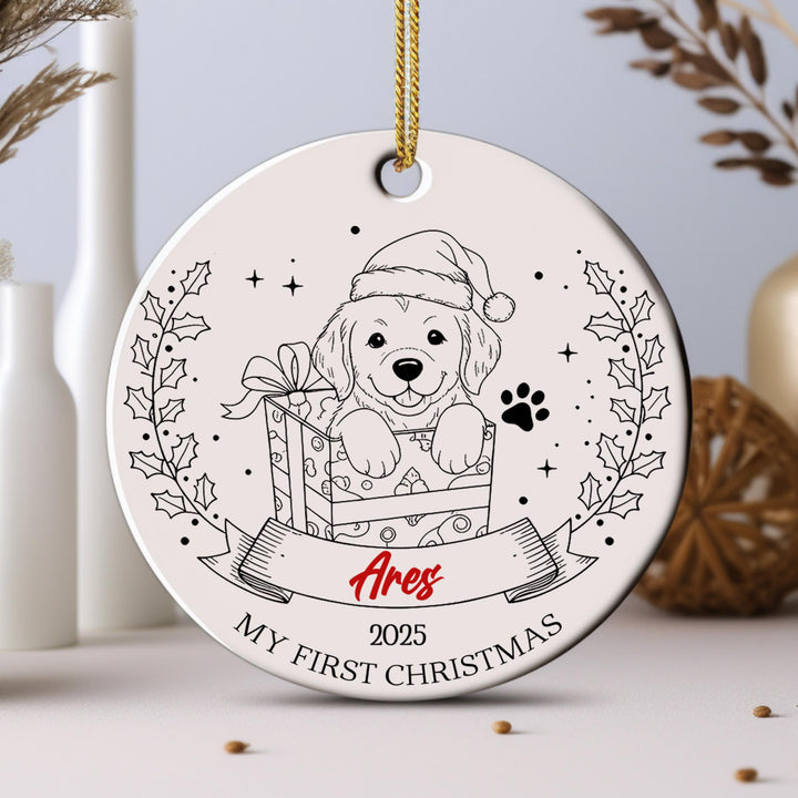 A white ornament with a black and white illustration of a dog wearing a Santa hat, holding a gift box, and surrounded by holly leaves and a ribbon.