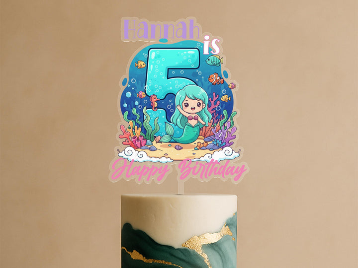 A birthday cake topper featuring a mermaid character and the number 5, with the text &quot;Hannah is 5&quot; and &quot;Happy Birthday&quot; written below.