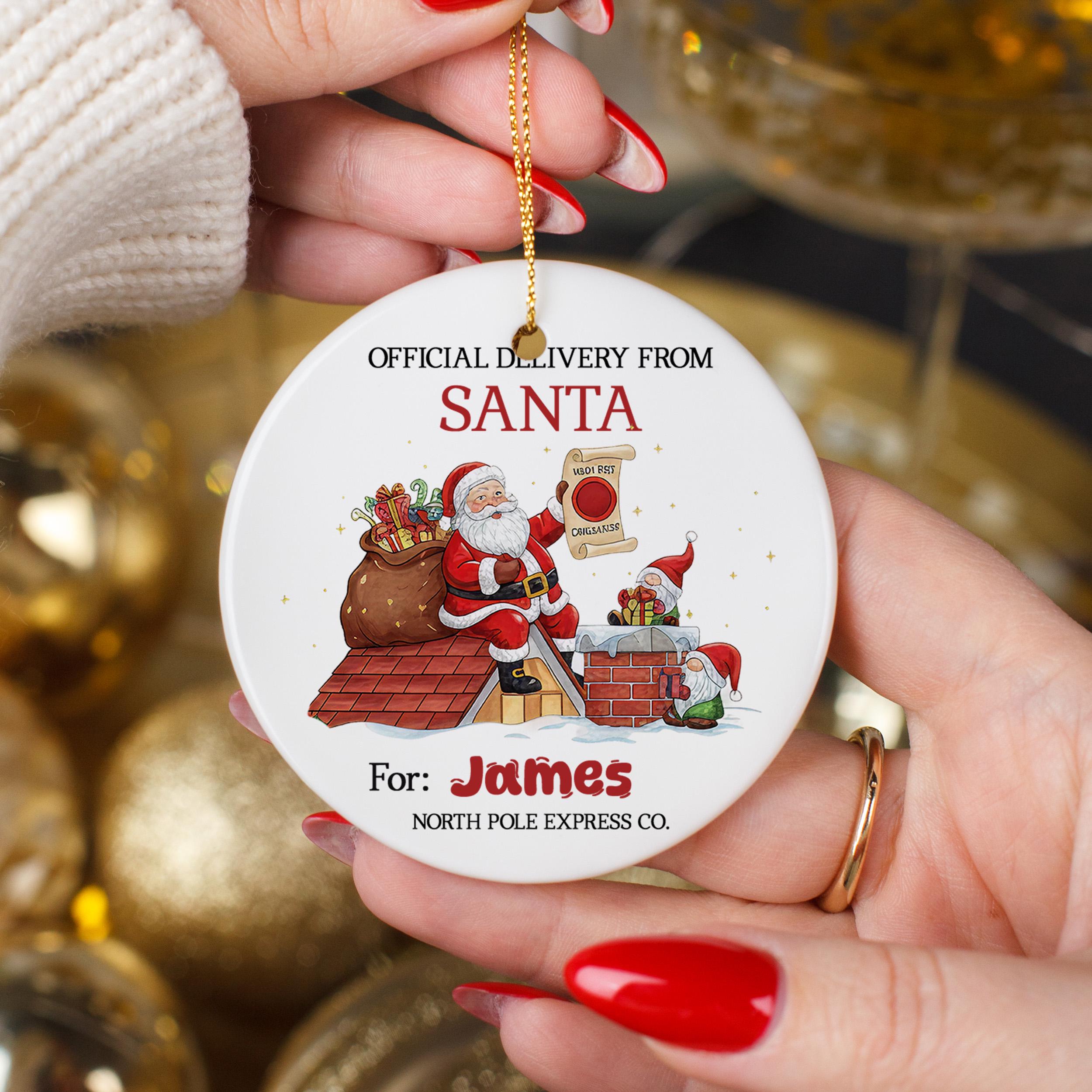 A person's hands holding a white ornament with a Santa Claus illustration and text, including "Official Delivery from Santa" and "For: James North Pole Express Co."