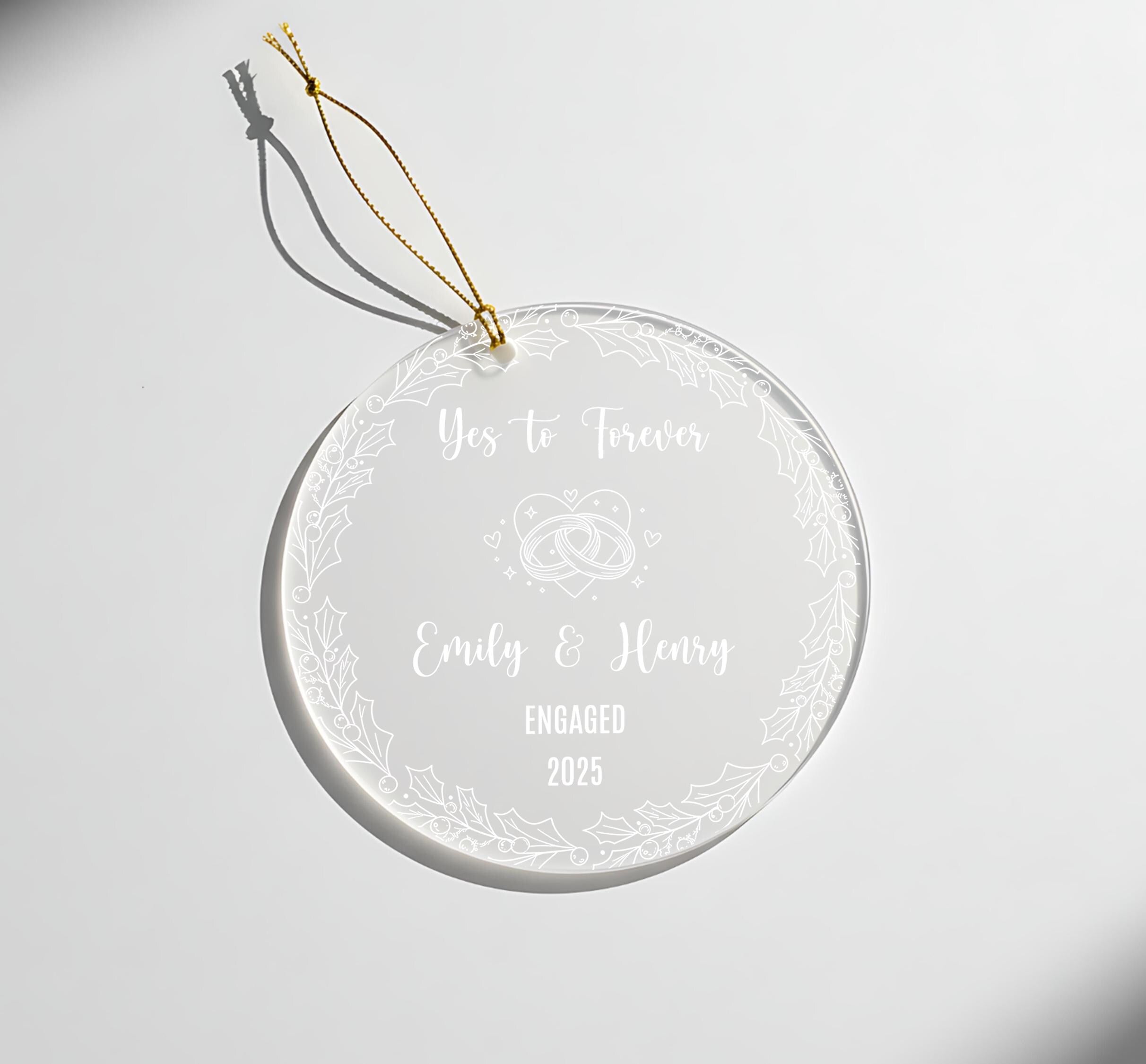 A clear glass ornament with a gold string attached, featuring a message that reads "Yes to Forever Emily & Henry Engaged 2025".