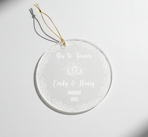 A clear glass ornament with a gold string attached, featuring a message that reads "Yes to Forever Emily & Henry Engaged 2025".