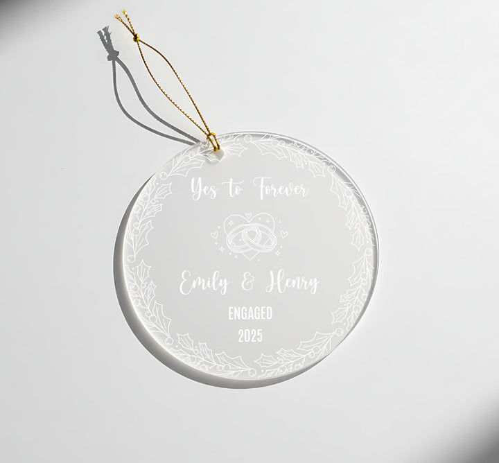A clear glass ornament with a gold string attached, featuring a message that reads &quot;Yes to Forever Emily & Henry Engaged 2025&quot;.