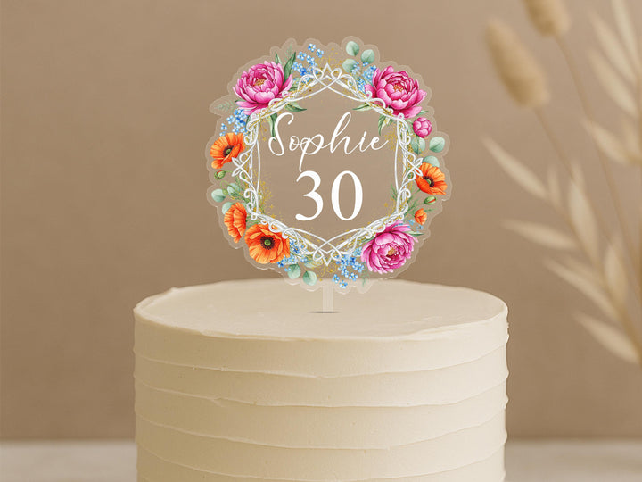A white cake with a floral wreath-shaped topper that says &quot;Sophie 30&quot; on it.