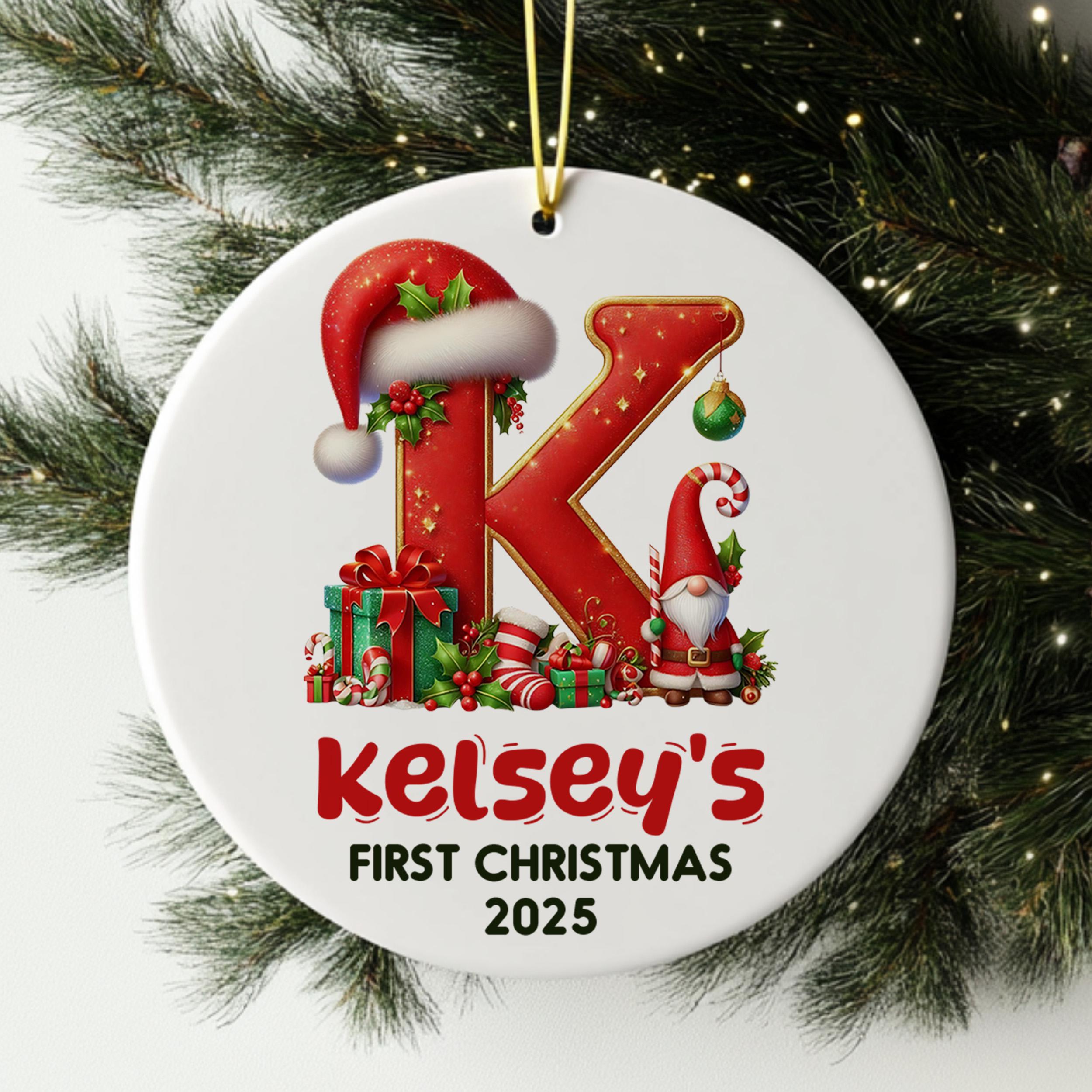 A white ornament with a red letter "K" and a gnome, along with various Christmas-themed elements such as presents and a ball. The ornament is hanging on a Christmas tree, and it is labeled as "Kelsey's First Christmas 2025".